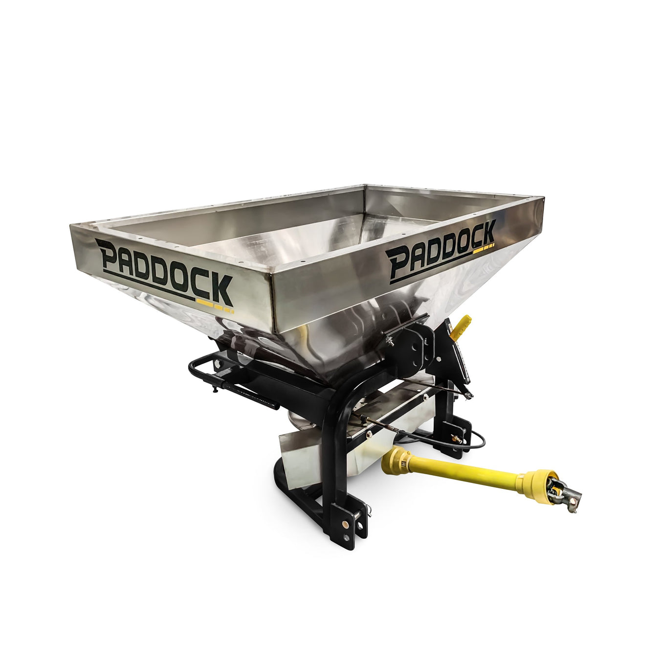 Stainless steel Paddock-brand fertilizer or seed spreader with a rectangular hopper, sturdy black frame, and a yellow PTO shaft attachment.