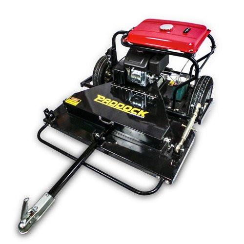 A heavy-duty Paddock tow-behind slasher mower with a red fuel tank, black metal body, two wheels, and a tow bar for attaching to vehicles.