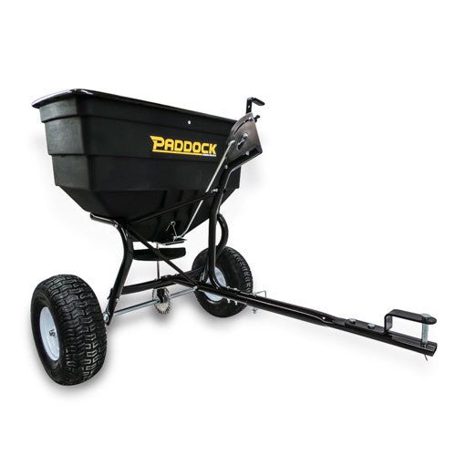 A black, heavy-duty Paddock tow-behind spreader with large wheels, a sturdy metal frame, and a long tow bar attachment for use with vehicles or tractors.