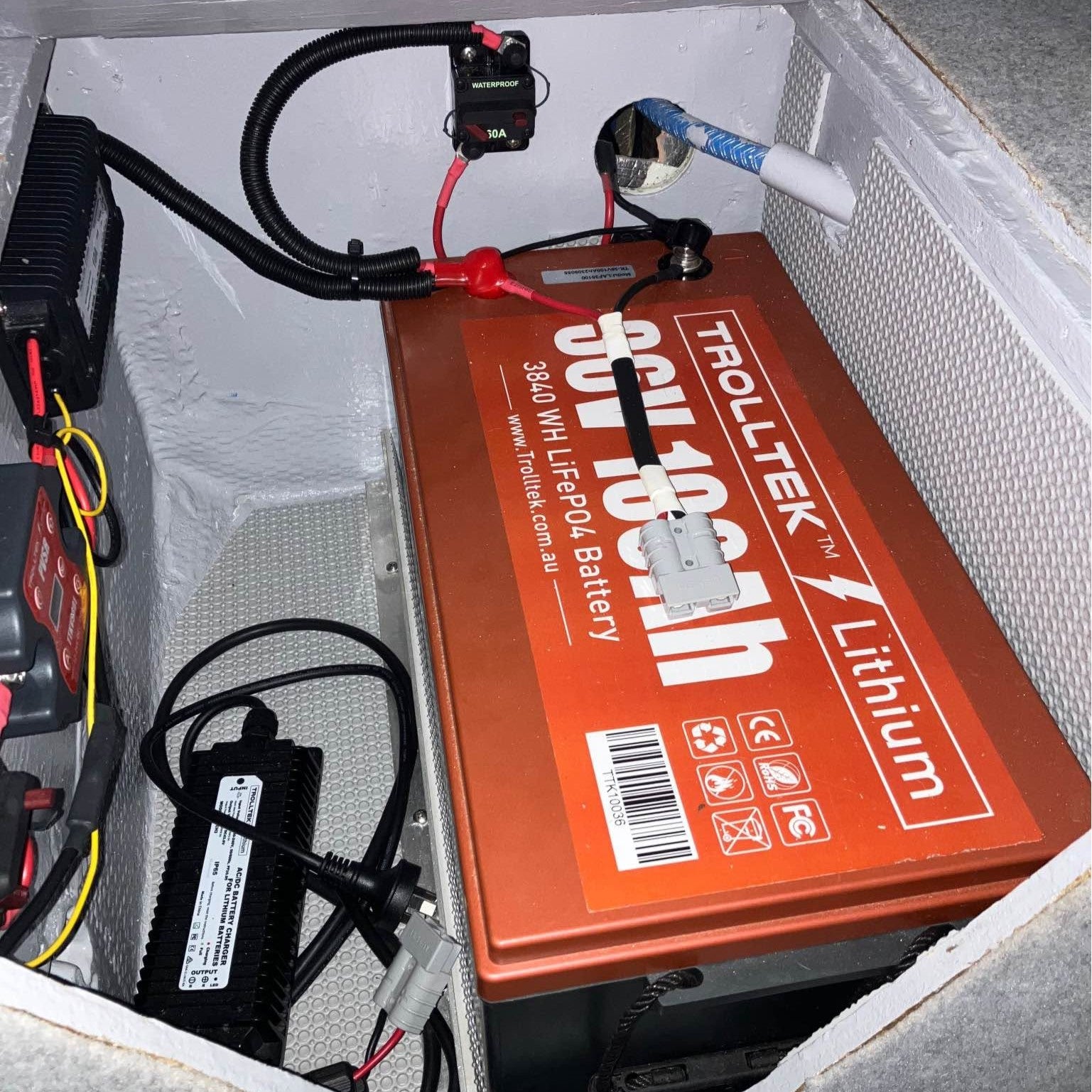 A large orange heavy-duty 36V Trolltek™ LiFePO4 Lithium Battery with Bluetooth, by Trolltek, is installed in a compartment with connected wires, a fuse, and a nearby battery charger. The setup features lithium batteries known for their long cycle life, all neatly organized inside a white enclosure.