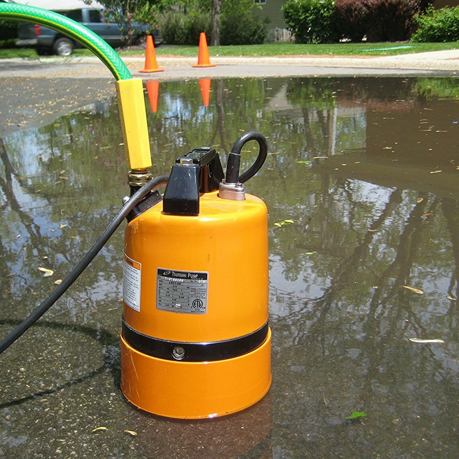 A Paddock Puddle Sucker Pump with attached hoses is placed in a large puddle on asphalt, efficiently removing standing water. Orange traffic cones and green shrubs are visible in the background.