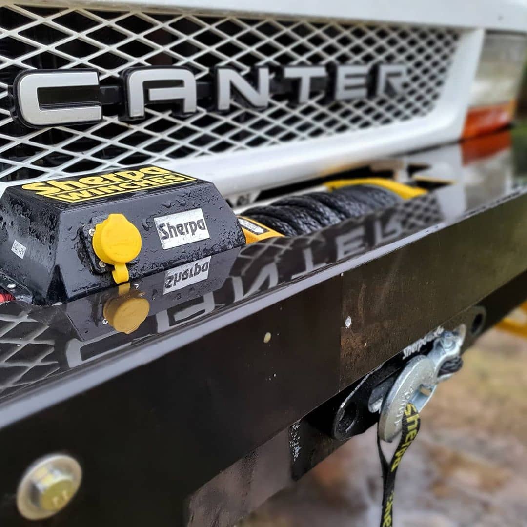 Close-up of a Sherpa 4x4 Black Edition ‘Stallion’ 25,000Lb Winch mounted on the front bumper of a white Canter truck, with a yellow hook and cable clearly visible.