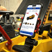 A smartphone, mounted inside construction equipment, displays a webpage featuring the Paddock™ Tracked Mini Loader Elite—a powerful 58hp Yanmar 2.1L 4-cylinder CRDi Turbo Diesel from Paddock—listed for sale. In the background, control levers and a blurred worker in safety gear emphasize the cutting-edge design and job site readiness of compact tracked loaders like this elite model for modern construction professionals.