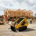 A Paddock Multi Hitch Jib Plate is fitted to a yellow compact track loader at a construction site with a wooden-framed house under construction. Construction workers and an orange excavator are also on site.