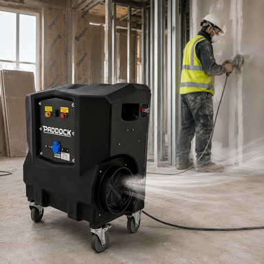 A construction worker sands a wall in a dusty, unfinished room with exposed framing and drywall. In the foreground, a Paddock Industrial Air Scrubber Filter from Paddock is running, efficiently filtering dust particles from the air with its advanced HEPA filtration system.