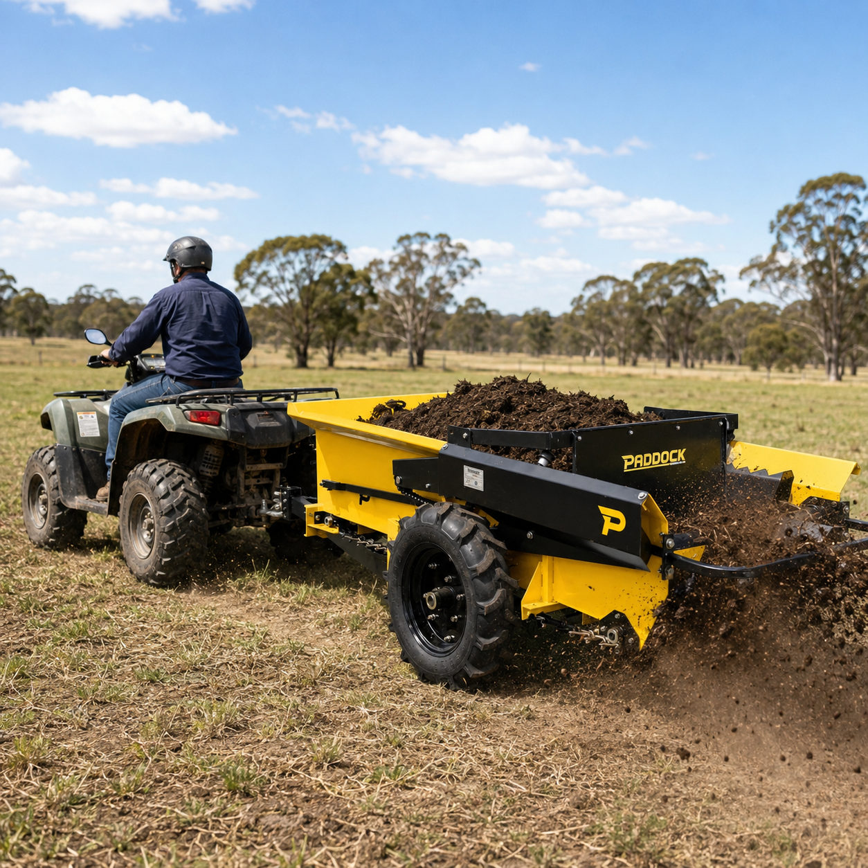 A person driving an ATV pulls a yellow Paddock Manure Spreader by Paddock, spreading soil or compost across a grassy field with trees and a blue sky in the background.