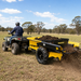 A person driving an ATV pulls a yellow Paddock Manure Spreader by Paddock, spreading soil or compost across a grassy field with trees and a blue sky in the background.