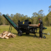 A person wearing ear protection feeds a log into a Paddock™ 15T Firewood Processing Machine, which uses an attached conveyor belt to stack split wood on the grass.