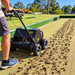 A person pushes a Paddock Motorised Lawn Aerator - Honda GX160 across a green turf surface, performing core aeration as it leaves small plugs of soil behind the machine.