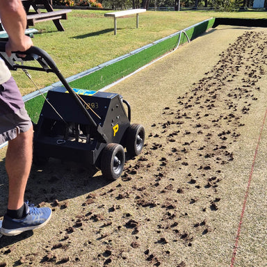 A person uses a Paddock Motorised Lawn Aerator powered by a Honda GX160 engine on a lawn bowling green, removing debris and leaving small clumps of soil on the grass. Benches and greenery are seen in the background.