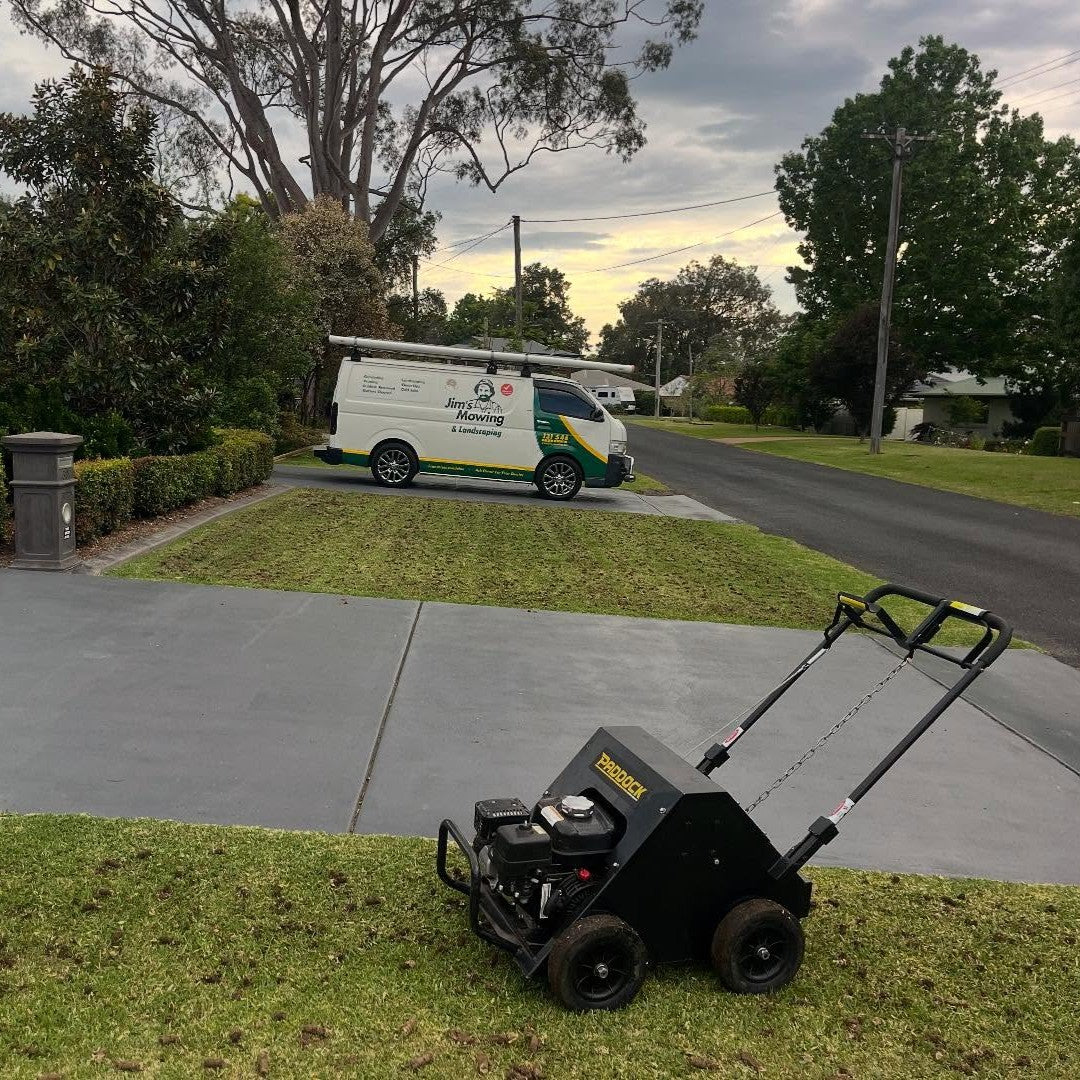 A Paddock Motorised Lawn Aerator - Honda GX160 sits on a grassy yard near a driveway. In the background, a white van with landscaping company signage is parked on the street, surrounded by trees and houses. This advanced lawn aerator from Paddock, powered by the dependable Honda GX160 engine, is perfect for maintaining healthy and vibrant lawns.
