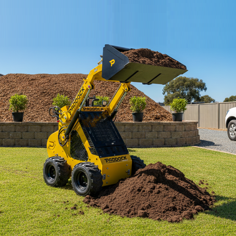 The Paddock Wheeled Mini Loader with a 23hp Honda Petrol Engine lifts soil onto a grass lawn. Behind the Paddock loader are a mulch pile, potted plants on a stone wall, and a white vehicle parked on gravel.