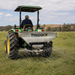 A person operates a green tractor equipped with a Paddock Tractor PTO Seed Fertiliser Spreader 600L, distributing materials onto a grassy field. The tractor features large tires and a metal attachment labeled “Paddock,” with hills and trees visible in the background.