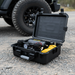 The Sherpa 4x4 'BOX AIR' Air Compressor (118 L/min) with yellow coiled hose and pressure gauge rests in its black case on gravel by an off-road vehicle's tire, ready for fast tyre inflation.