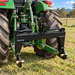 A Paddock™ 3-Point Quick Hitch – Cat I & Cat II from Paddock, finished in black, is attached to the rear of a green tractor. The setup is situated on grassy ground in a scenic rural outdoor area with trees and a fence in the background. This essential piece of Paddock tractor implement equipment enables fast and efficient connections for a variety of tasks around your property.