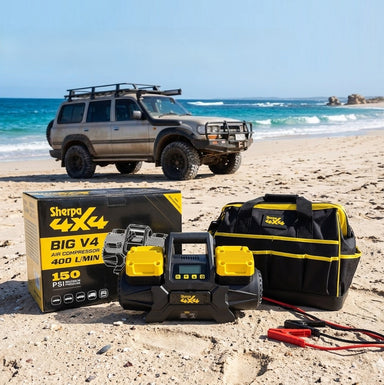 A Sherpa 4x4 'BIG V4' Air Compressor (400 L/min) with Pre-set Pressure Control System, its box, and a tool bag sit on a sandy beach with a 4WD nearby—ideal for tyre inflation and portable air needs under sunny skies.