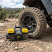 The Sherpa 4x4 'BIG V4' Air Compressor (400 L/min) with Pre-set Pressure Control System sits on rocky ground by a muddy off-road tire, ready for fast tyre inflation in tough outdoor conditions.