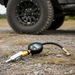 A Scintex Sherpa 4x4 Digital Inflator Nozzle with Nitto fittings and a round display rests on gravel by an off-road vehicle tire. The battery-compatible gauge is housed in a tough black rubber case.