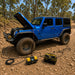 A blue Jeep with its hood open is parked on a rocky dirt road in a forest. Jumper cables connect a battery charger and a tool bag to the vehicle, while the Sherpa 4x4 ‘BIG V4’ Quad-Air Bundle – (400 L/min) 4-Tyre Inflation System from Sherpa 4x4 is ready nearby for quick 4x4 tyre inflation.