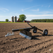 The Paddock ATV & Quad Bike Blade by Paddock, featuring wheels and a blade, sits on freshly tilled soil in a field. Behind it are two silos, a red barn, green trees, and a blue sky with clouds in a rural landscape.