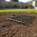 A Paddock Drag Harrow by Paddock is placed on green grass in a backyard, with garden beds, a wooden fence, and a house visible in the sunny background.