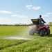 A person uses the Paddock 12V ATV Seed and Fertiliser Spreader by Paddock to distribute fertilizer across a large grassy field under a clear blue sky.