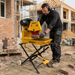 A man wearing protective gear operates a Paddock Brick Table Saw, equipped with a powerful Honda engine, to cut bricks at a construction site surrounded by unfinished buildings and stacks of bricks in the background.