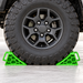 A large off-road vehicle tire secured by a Sherpa 4x4 Wheel Chock Set, featuring two bright green heavy-duty wheel chocks.