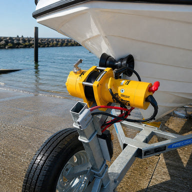 A Sherpa 4x4 ‘Grunter’ Electric Boat Trailer Winch is mounted on a boat trailer near the front of a white boat at a boat ramp by the water, with a rocky seawall in the background.