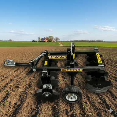 A Paddock Disc Harrow by Paddock sits on a freshly plowed field, with a farmhouse, silo, and trees visible in the background beneath a clear blue sky.