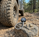 The Sherpa 4x4 Digital Tyre Deflator & Gauge with Quick Connect Fittings rests on a rock beside a muddy off-road tyre along a rugged, rocky forest trail.