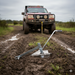 The Sherpa 4x4 Ground Anchor from Sherpa 4x4 is secured in muddy ground on a grassy dirt track, with a cable connected to a 4x4 vehicle ready for recovery under an overcast sky.