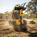 A Paddock Post Hole Digger Attachment by Paddock is used with a yellow skid steer to drill into the soil at a sunny construction site, as workers in vests and helmets work nearby in a fenced yard with wooden posts and trees.