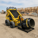 A yellow Paddock skid steer fitted with a Paddock Cement Mixer Attachment is parked on a construction site amid wooden frames and gravel piles.