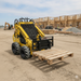 A Paddock Mini Loader, equipped with Paddock Mini Loader Forks, lifts a wooden pallet at a construction site, with unfinished wooden structures and building materials visible in the background.