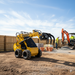 A Paddock mini loader with a Paddock Ripper Attachment moves material bags at a construction site, while workers and an orange excavator operate nearby, all within a wooden fence under a clear blue sky.