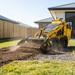 A Paddock Levelling Bar Attachment by Paddock levels soil on a gravel path in a modern backyard, featuring green grass, wooden fencing, and a white house in the background.