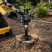A Paddock 350mm (14″) Stump Planer Attachment from Paddock, mounted on a yellow skid steer, efficiently grinds away a tree stump in a landscaped outdoor area as wood chips are thrown by the heavy-duty planer in action.