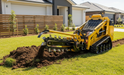 A Paddock Trencher Attachment by Paddock digs a narrow trench in the grass of a residential yard, with modern houses and landscaped gardens visible in the background.