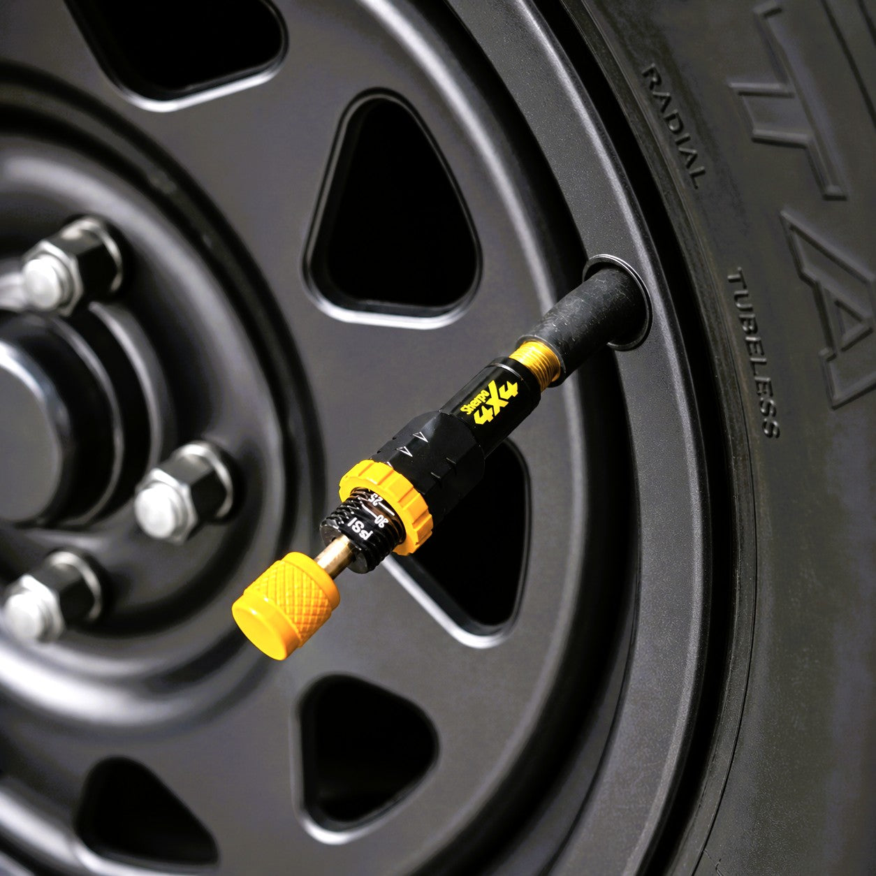 Close-up of a Sherpa 4x4 Tyre Deflators - 4pk unit attached to a vehicle’s valve stem, showing the yellow and black design and rusty metal wheel with lug nuts in the background.