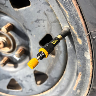 Close-up of a Sherpa 4x4 Tyre Deflators - 4pk unit attached to a vehicle’s valve stem, showing the yellow and black design and rusty metal wheel with lug nuts in the background.