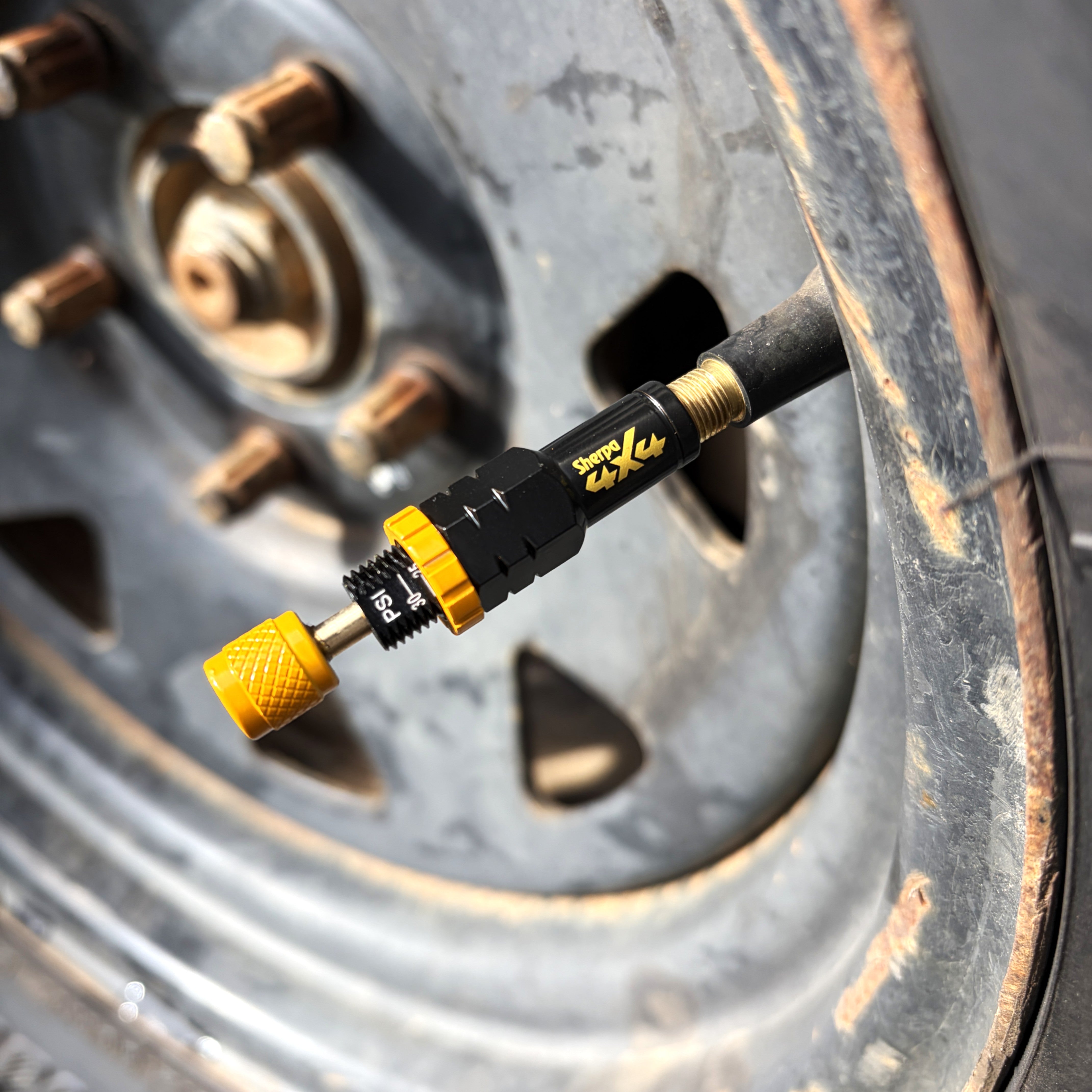 A Sherpa 4x4 Tyre Deflators - 4pk, in black and yellow, is attached to a car tire’s valve stem, with the tire and a rusty wheel rim from Sherpa 4x4 visible in the background.