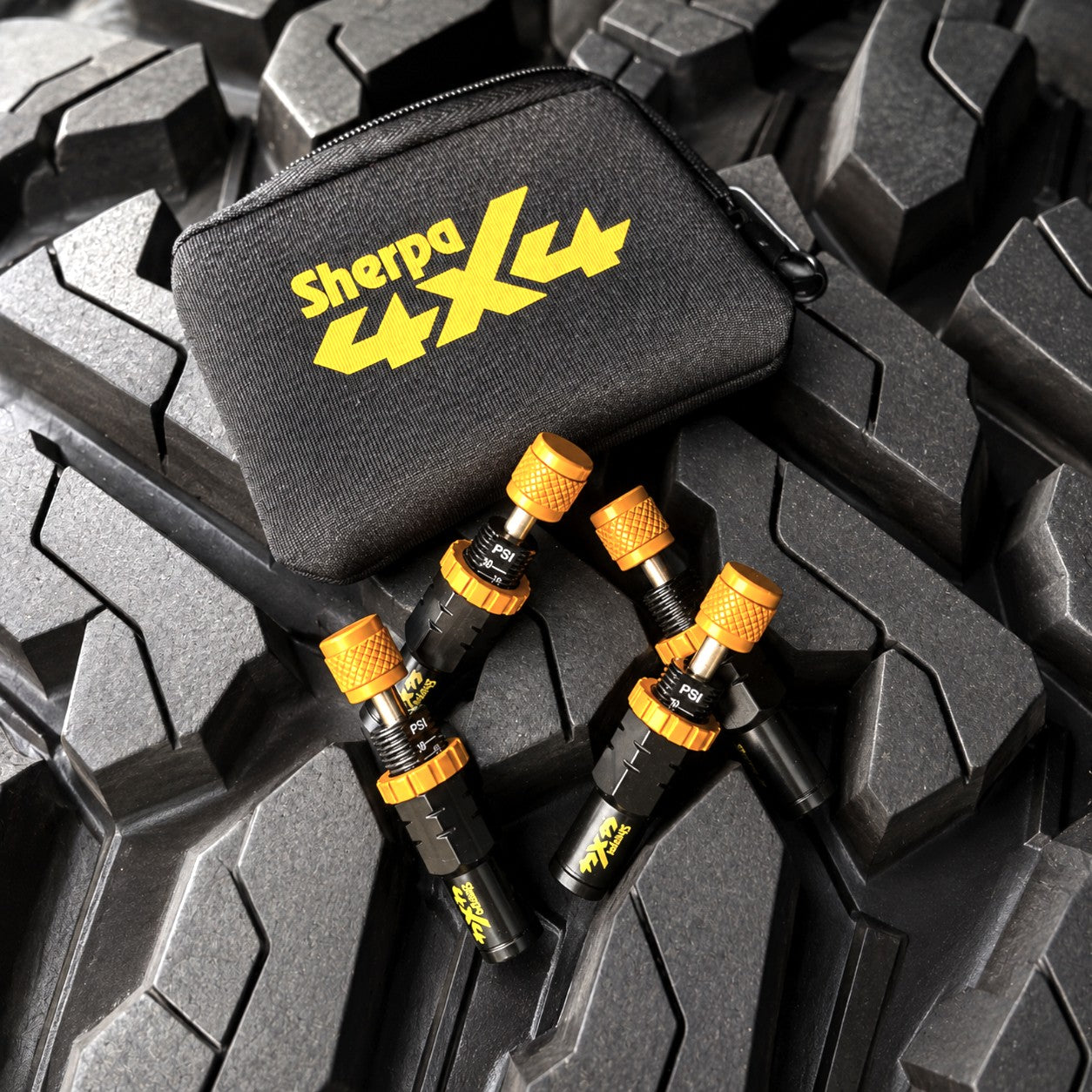 Four Sherpa 4x4 Tyre Deflators and a black case sit on the tread of a large, wet off-road tire—fast, reliable tyre deflation tools from Sherpa 4x4, perfect for any off-road adventure.