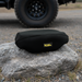 A Sherpa 4x4 Neoprene Winch Cover rests on a large gray rock outdoors, with a blurred off-road vehicle tire on gravel in the background, emphasizing rugged protection for your winch.