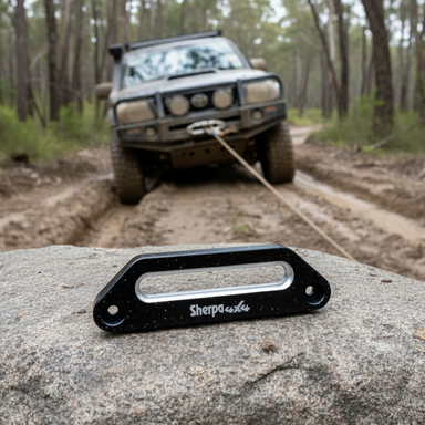 A Sherpa 4x4 Winch Hawse Fairlead by Sherpa 4x4 is attached to a rock in the foreground, while a muddy off-road vehicle with a synthetic rope winch is being recovered along a forest dirt trail in the background.