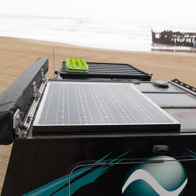 A close-up of Enerdrive Fixed Mono Solar Panels with a black frame mounted on a vehicle parked on a sandy beach, set against the ocean and a rusted shipwreck.