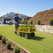 Wearing a sun hat, a person pulls the Paddock Electric Power Wheel Cart - 48V by Paddock with plants across a tidy lawn. Nearby are potted plants, mulch, and a modern house with solar panels under a clear sky.