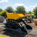 A Paddock Tracked Lifting/Tipping Power Barrow - 800kg Capacity is parked on a construction site, as two workers in safety vests and helmets work near a wheelbarrow and stacked bricks. Trees and a clear sky can be seen in the background.