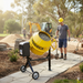 A construction worker in a hard hat and gloves walks near a yellow Paddock Cement Concrete Mixer 160L by Paddock with an electric motor on a paved path, while an excavator and building materials are seen in the background at a construction site.
