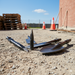 Paddock Jack Hammer Attachment Tools are scattered on gravel at a construction site, surrounded by traffic cones, wooden pallets, and a brick wall under a clear blue sky.